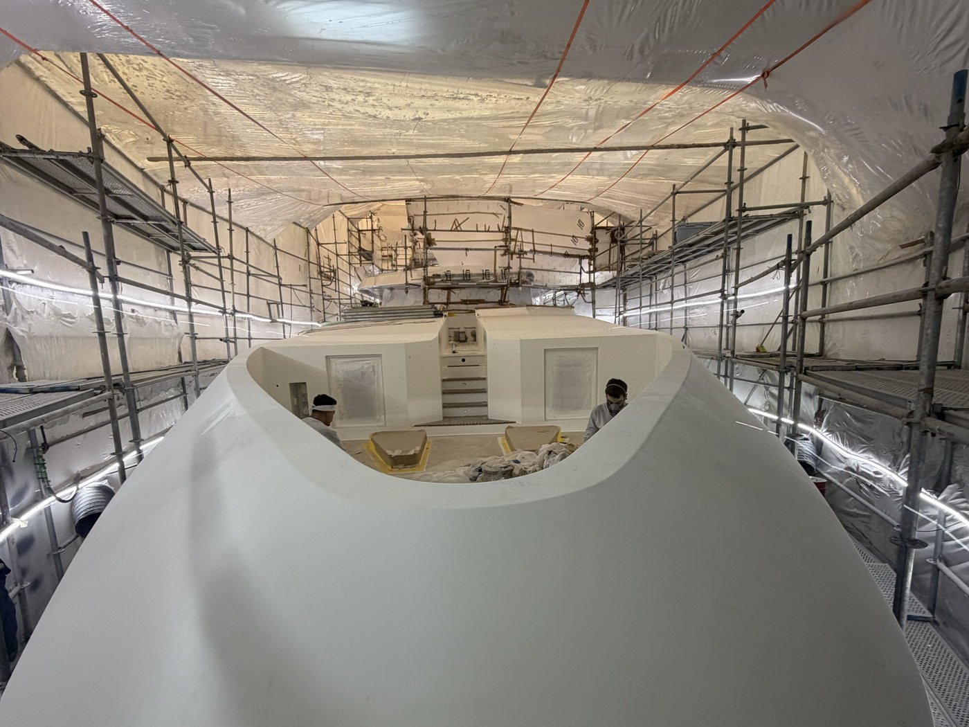 A yacht hull seen bow-on inside a tented paint hall, surrounded by scaffolding during a refit