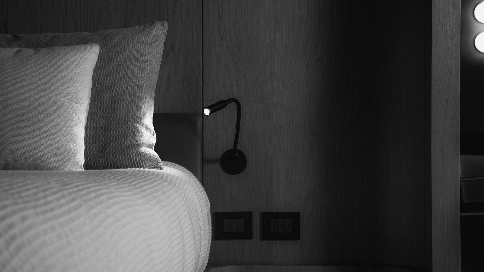 A black and white cabin corner: a made-up bed, a plain pillow behind a coarse woven scatter cushion over a ribbed throw in parallel folds, a flat straight-grain timber panel with one flush vertical joint, a bedside shelf with a radiused corner at lower right, and a black gooseneck light burning above two flush black plates.