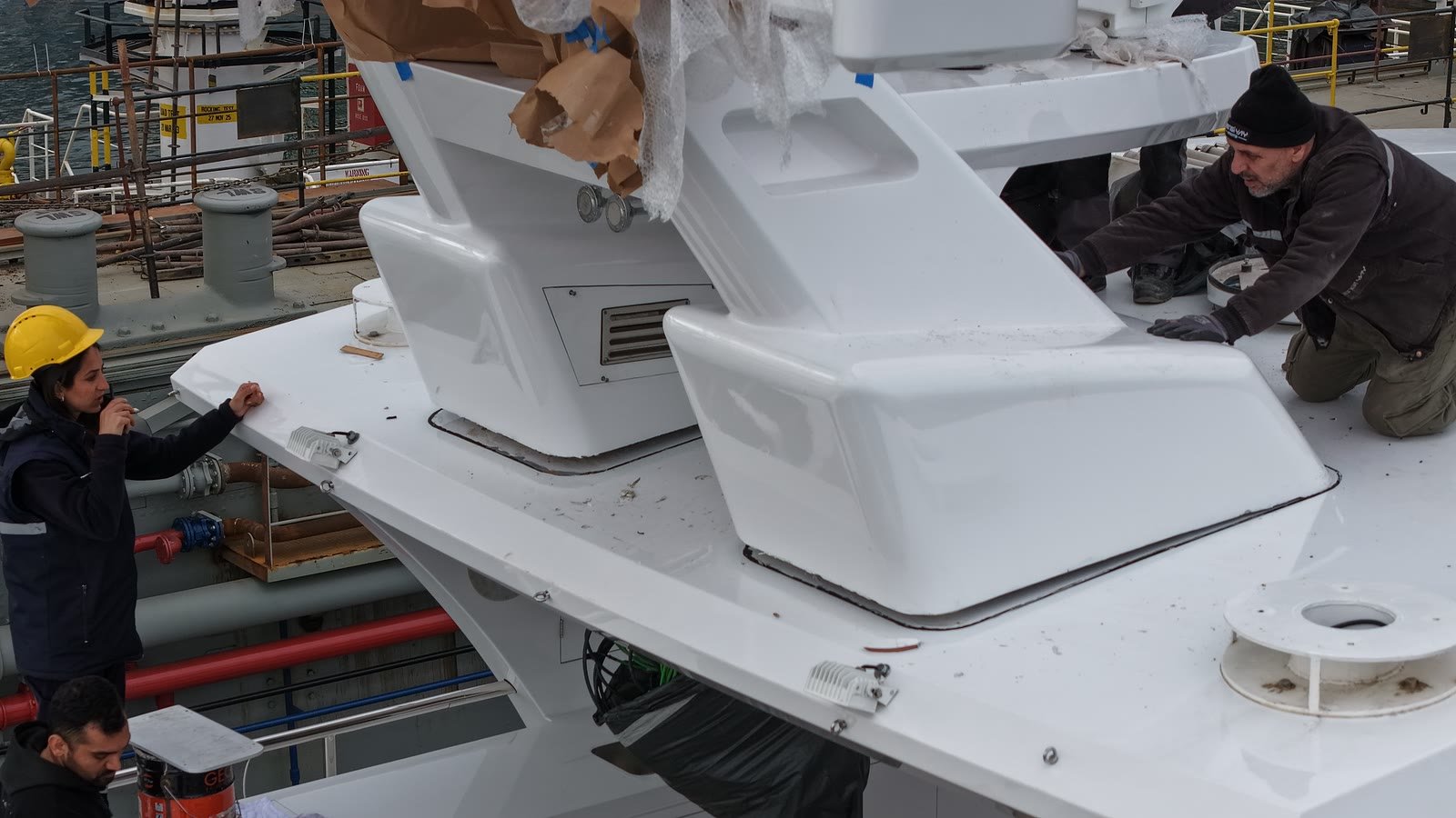 Two men working on a newly built white flybridge structure on a yacht in the yard, one in a yellow hard hat steadying the edge while the other kneels at the moulding, protective wrapping still on the mast section