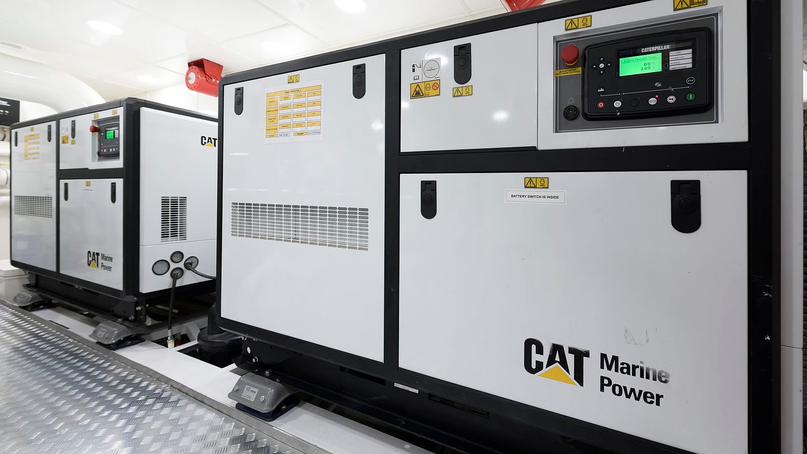 Two CAT Marine Power generator sets in a white machinery space, standing on resilient feet on chequer-plate flooring; the nearer enclosure carries a Caterpillar control panel reading engine coolant temperature, a printed maintenance schedule taped to the door, hazard decals and a notice reading battery switch is inside.