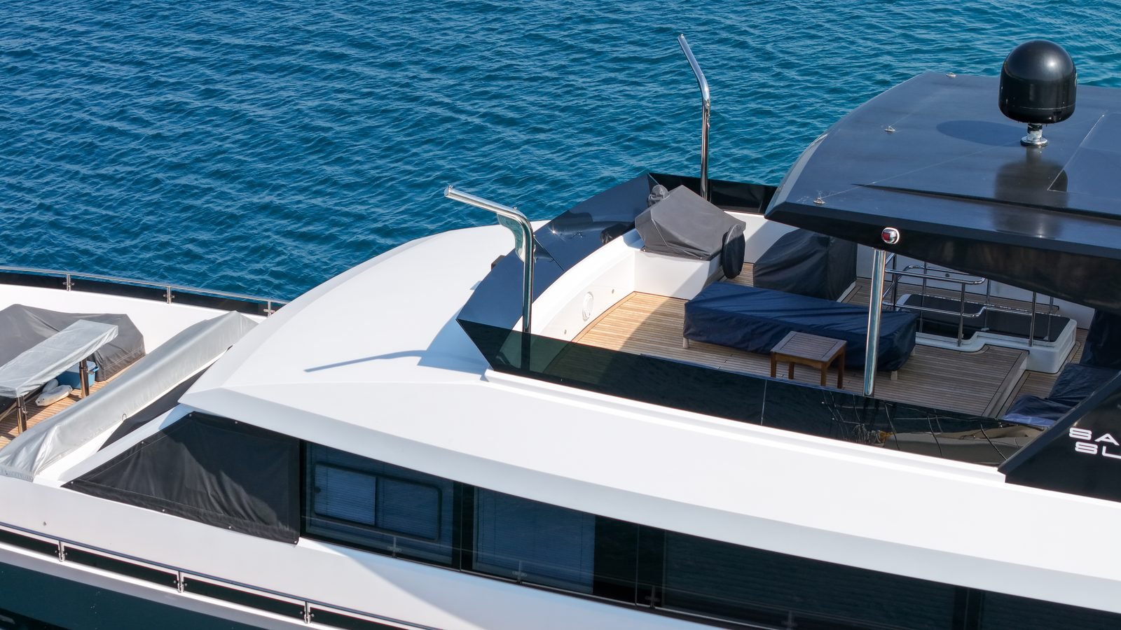 A motor yacht's upper deck seen from above and astern: teak decking, a sunpad under a fitted navy cover, two polished davit arms and a black satellite dome on the hardtop, with a covered tender on the side deck below