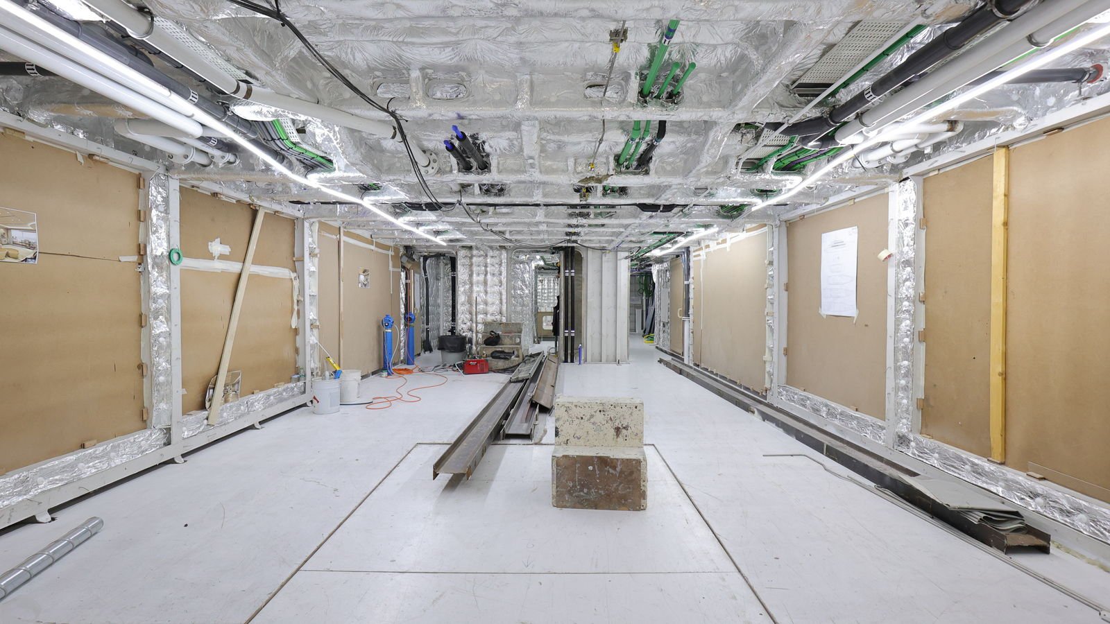 A stripped interior corridor of a yacht under refit, bare framing and foil insulation exposed, temporary lighting and cable runs overhead and a steel section lying on the sole