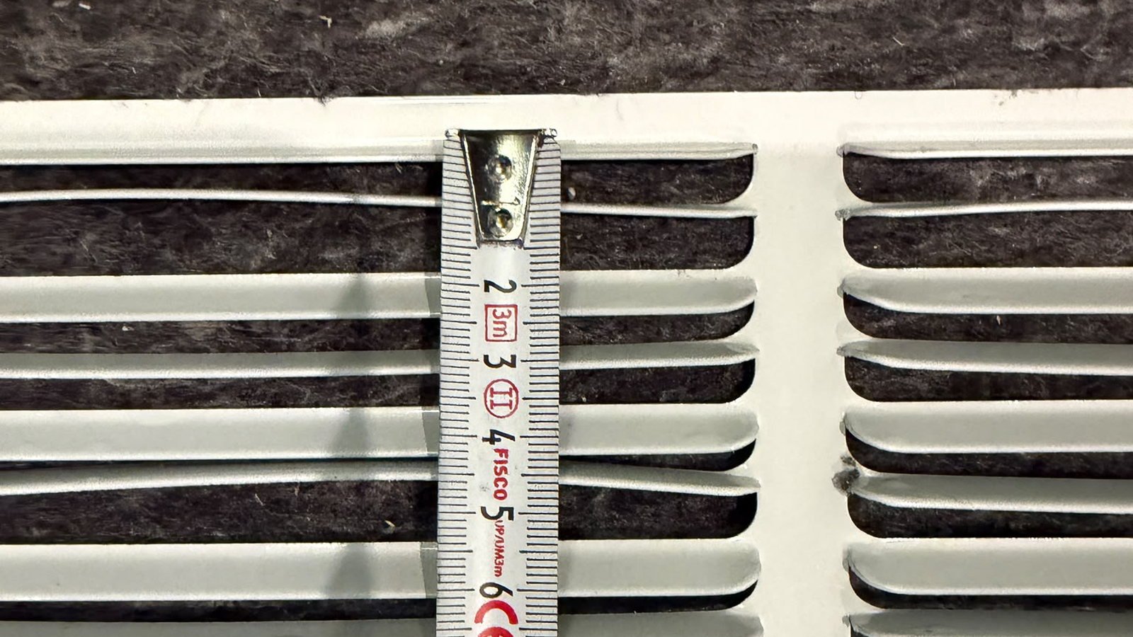A dimension being taken off a cream-white louvred panel, shot square-on: a steel tape is hooked over the panel's top edge just left of the plain vertical stile, and its blade runs vertically down across the horizontal slats, with the numerals two to six and the scale markings legible. A dark grey fibrous lining shows behind the slats, and the panel's top rail is chipped and marked in places.