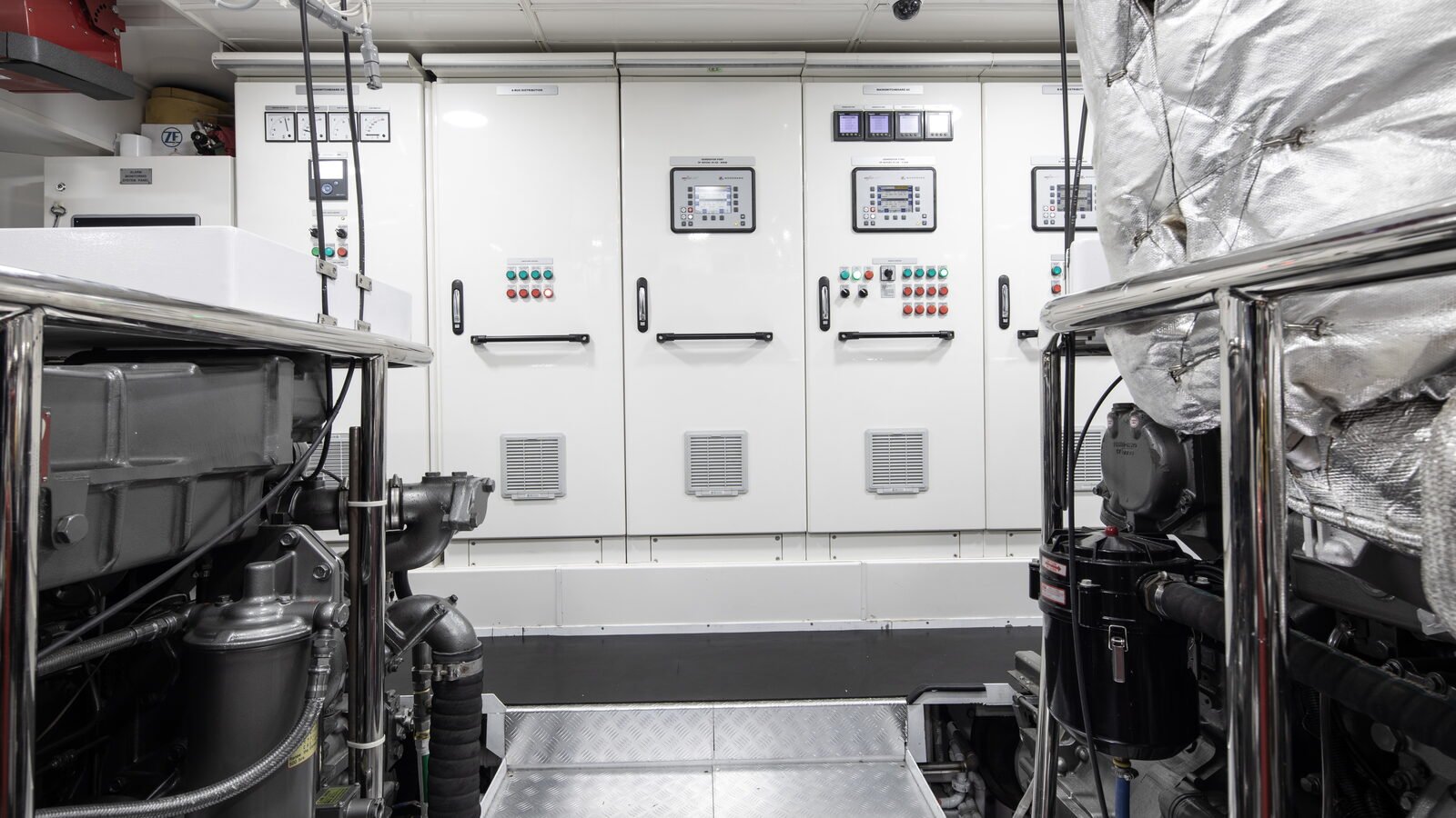 The main switchboard of a yacht seen from between her two main engines. Five white cubicle doors stand in a row across the frame, each with a long black D-handle set at the same height, and a small printed legend plate above it. Three of the doors carry a control module with a screen and a keypad set into a dark surround; one carries a row of round analogue meters across the top; two carry banks of small green and red indicator lamps, one of them with a black selector switch beside them. A square louvred ventilation panel is let into the lower part of three doors. A continuous white plinth runs along the foot of the cubicles above a dark deck. In the foreground, port and starboard, the tops of the two main engines fill the lower corners: grey castings, black spin-on filters, braided flexible hose and black pipework, with a polished stainless handrail standing in front of each. A silver foil-lagged exhaust run fills the right-hand edge of the frame from top to bottom. A chequer-plate step leads down the centreline between the engines towards the switchboard.