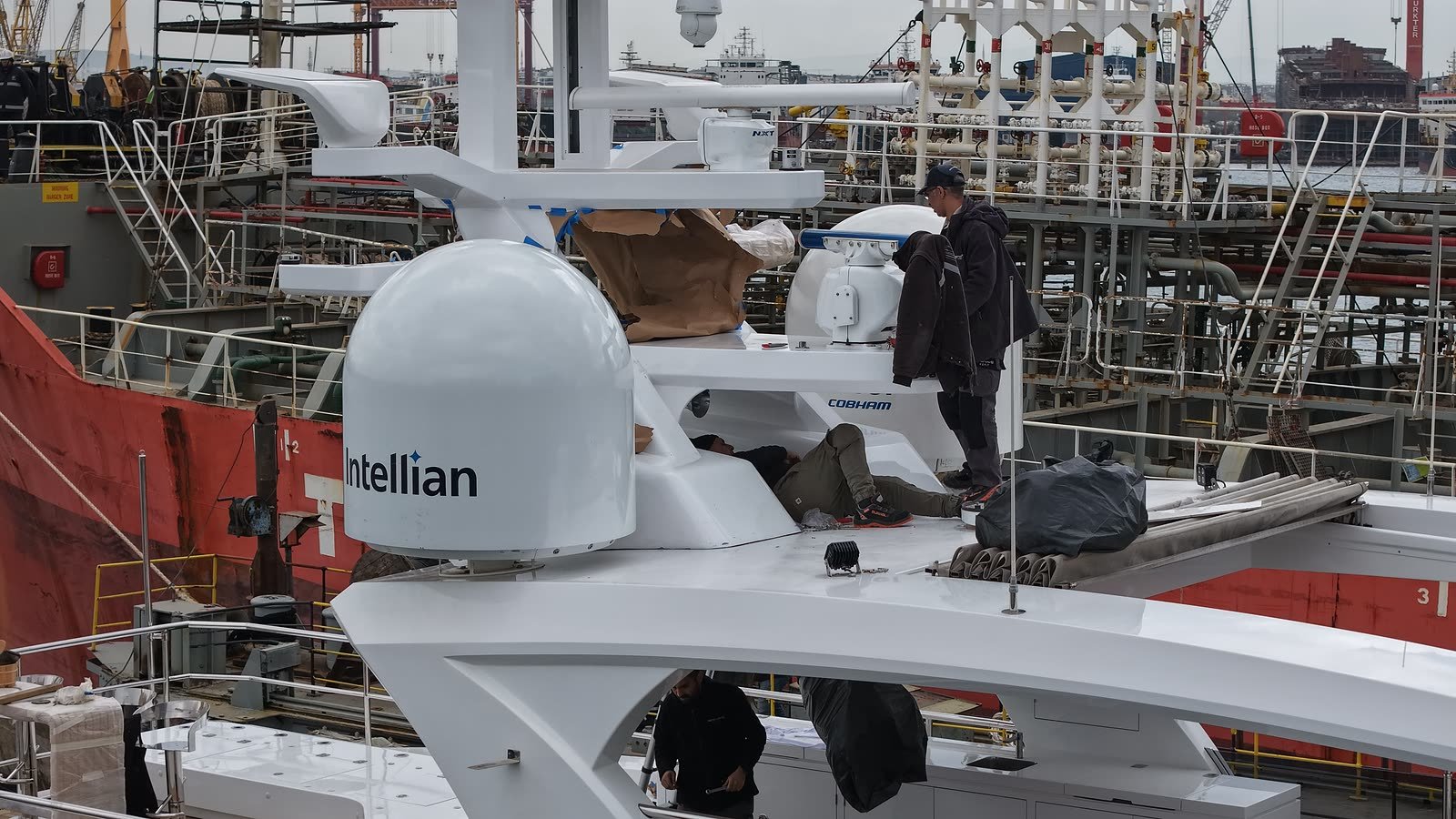 A satellite communications dome and mast-mounted electronics being fitted on a yacht's flybridge, a fitter working at the mast with the red hull of a commercial ship alongside