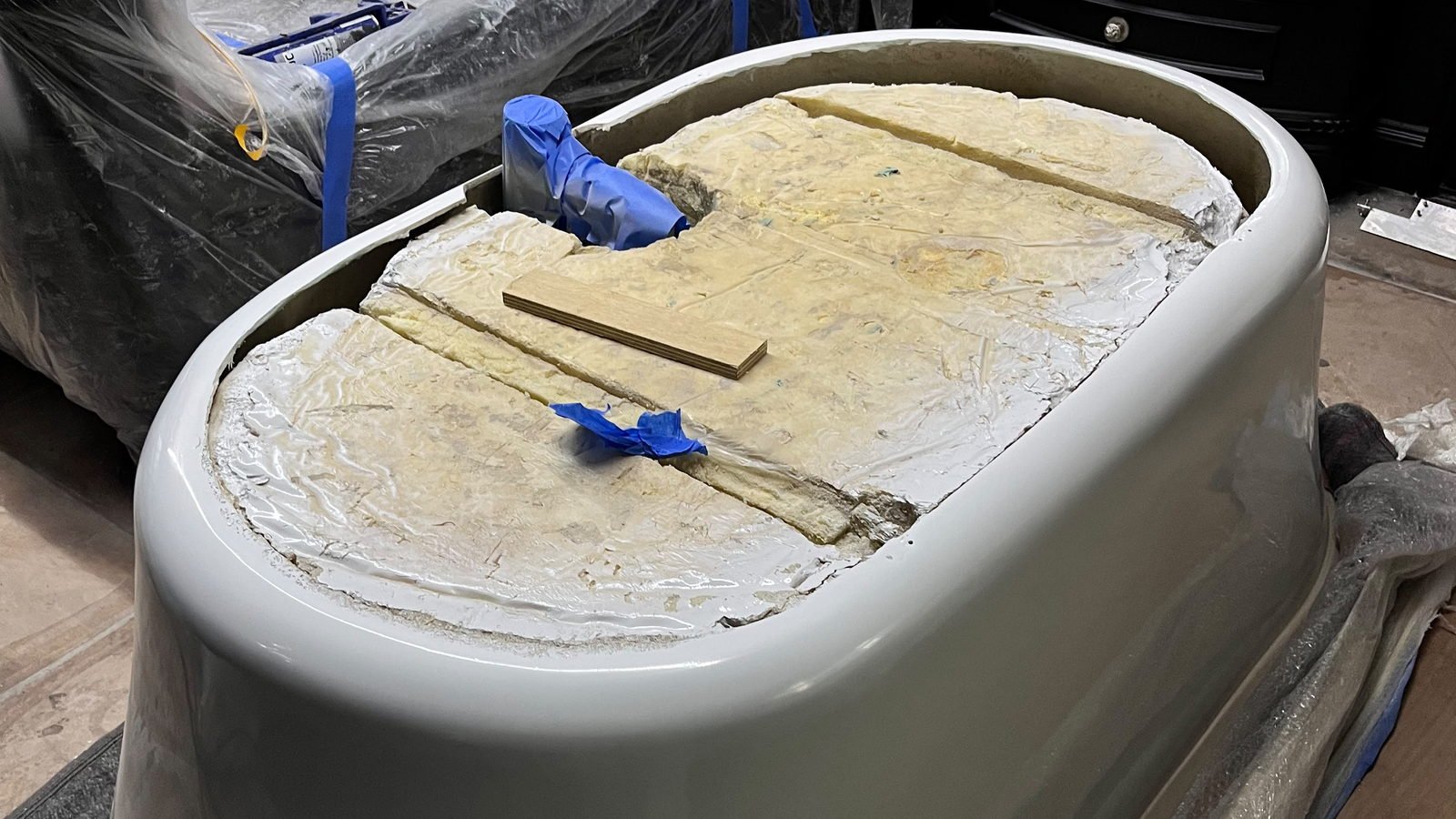 A large moulded bath shell in glossy cream-grey composite, out of the vessel and standing upside down on protective sheeting on a boarded workshop floor, its exposed underside showing pale yellow batt insulation laid in strips, ragged white bonding lines around the rim, two strips of blue masking tape and a loose plywood offcut, with wrapped panels and a dark cabinet behind it and nobody in the frame.