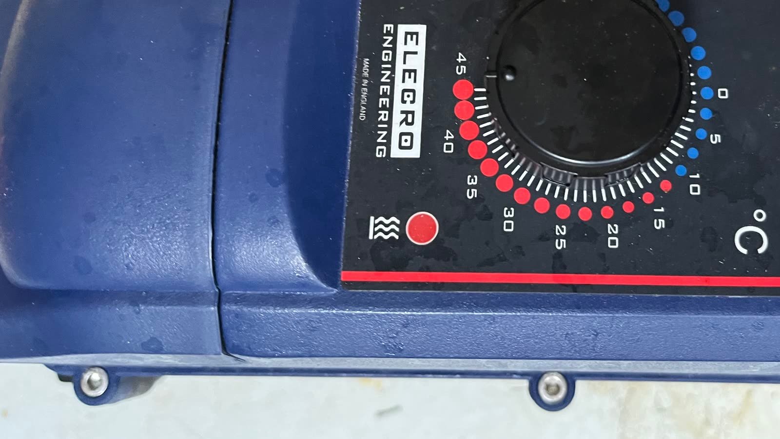A close view down onto the same unit's dark blue moulded casing and the black control face set into it. A large plain rotary knob sits at the centre of a graduated scale running from an off position through a row of blue dots at the low end to red dots at the high end, with the numbers stepping round it in degrees Celsius. Two round indicator lamps sit beside the dial, one amber and one red, and a fine red line runs along the top and the bottom edge of the face. Two stainless screws hold the lower edge of the casing, and a lighter grey surface shows beyond it at the bottom of the frame.