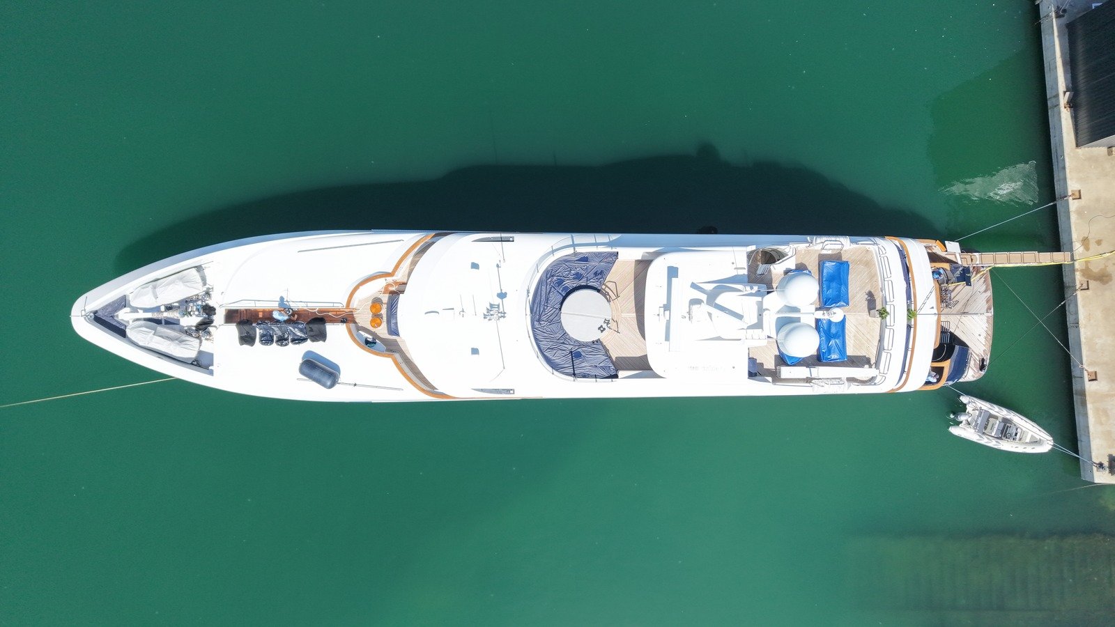 A motor yacht photographed vertically from directly above, moored alongside a concrete quay with mooring lines and a gangway ashore, teak side decks, blue sunpad covers, two white satcom domes, deck gear under covers forward and a RIB tender made fast aft.