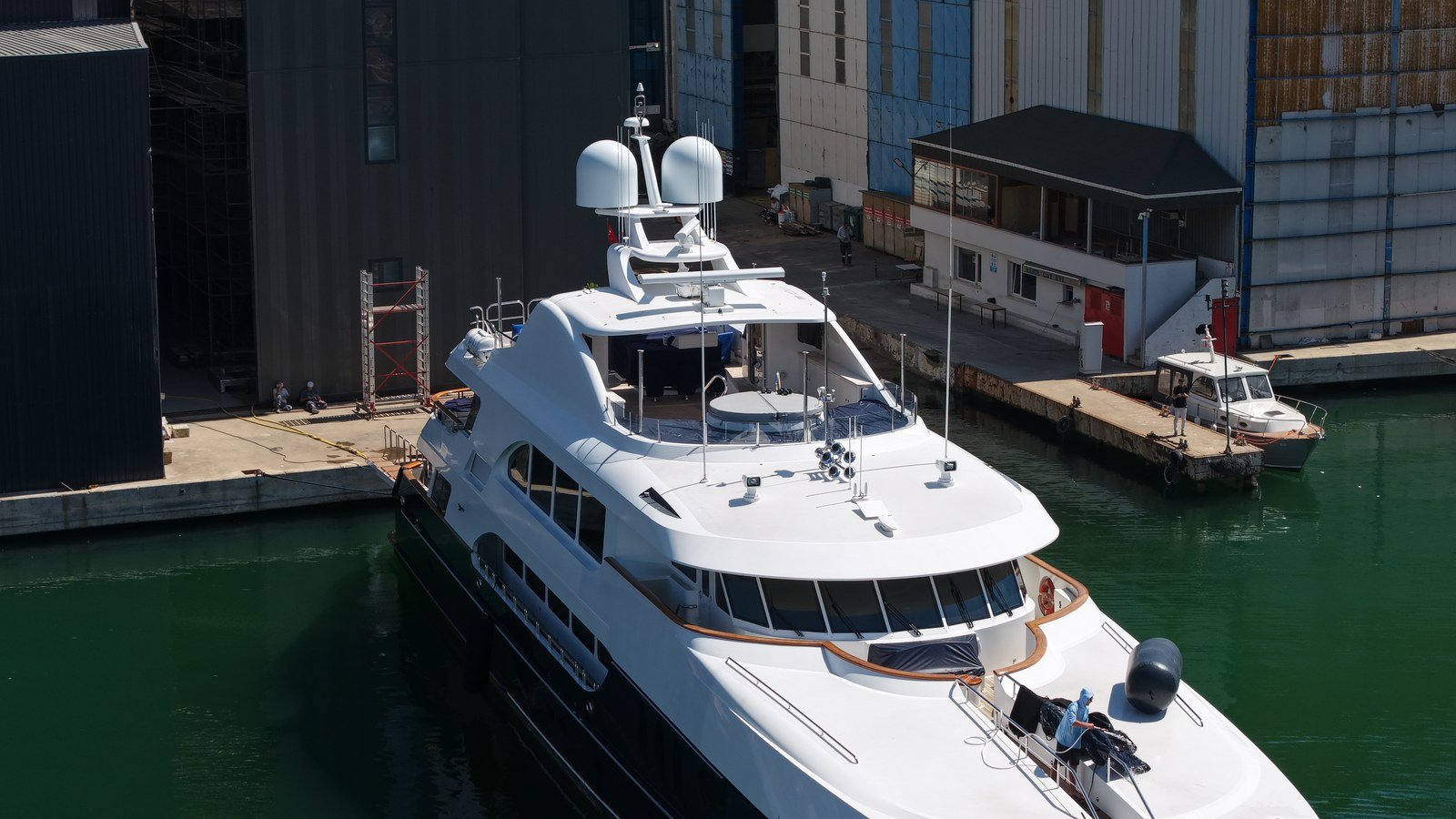 A white superyacht with a dark blue hull lying alongside the yard quay at Tuzla, a man in a blue shirt working at the bow rail, a red-and-white mobile scaffold tower and two people on the quay beside her.