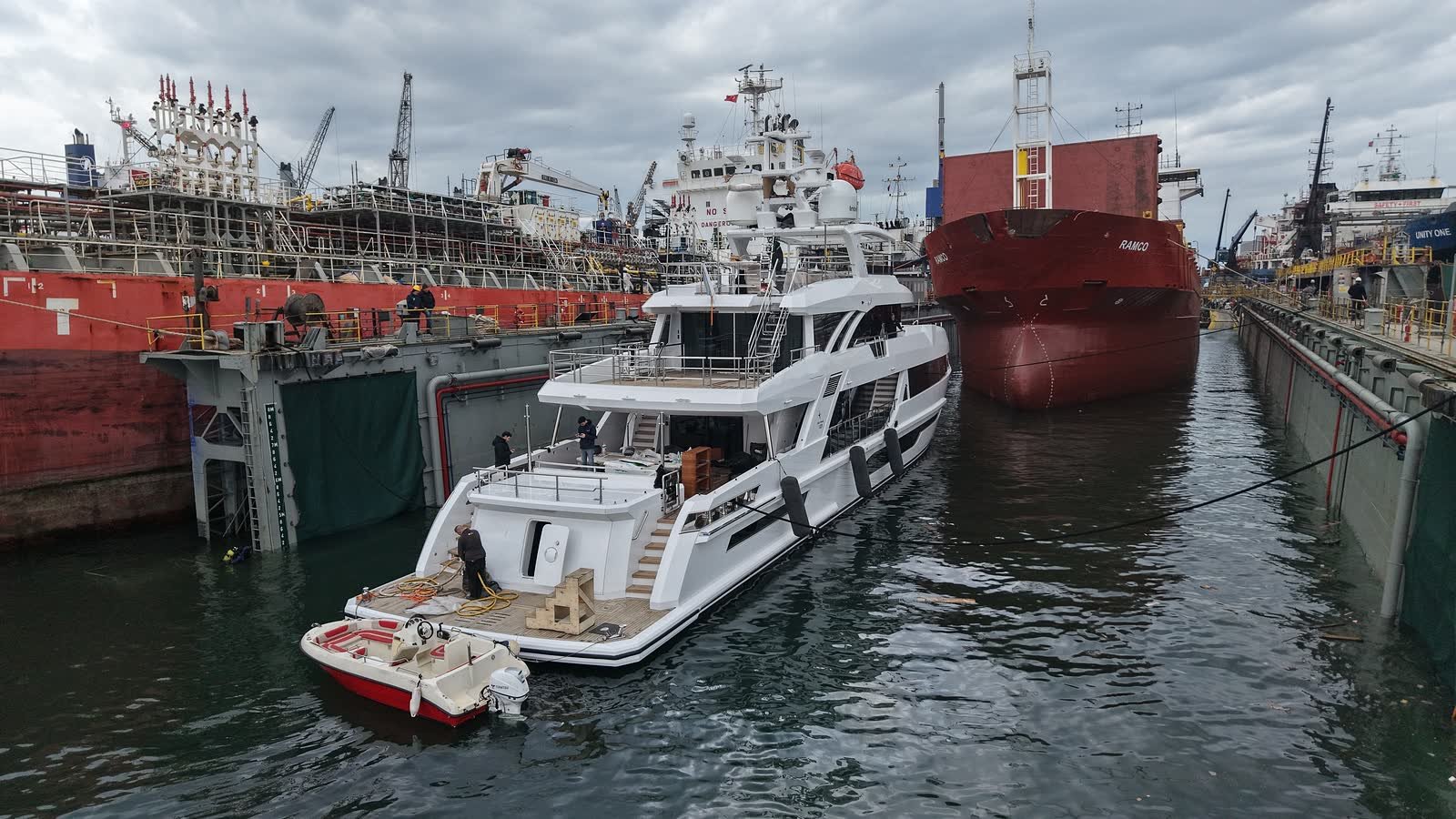 A motor yacht afloat inside a floating dock during a refit, a workboat alongside her and a commercial hull ahead, dock walls either side
