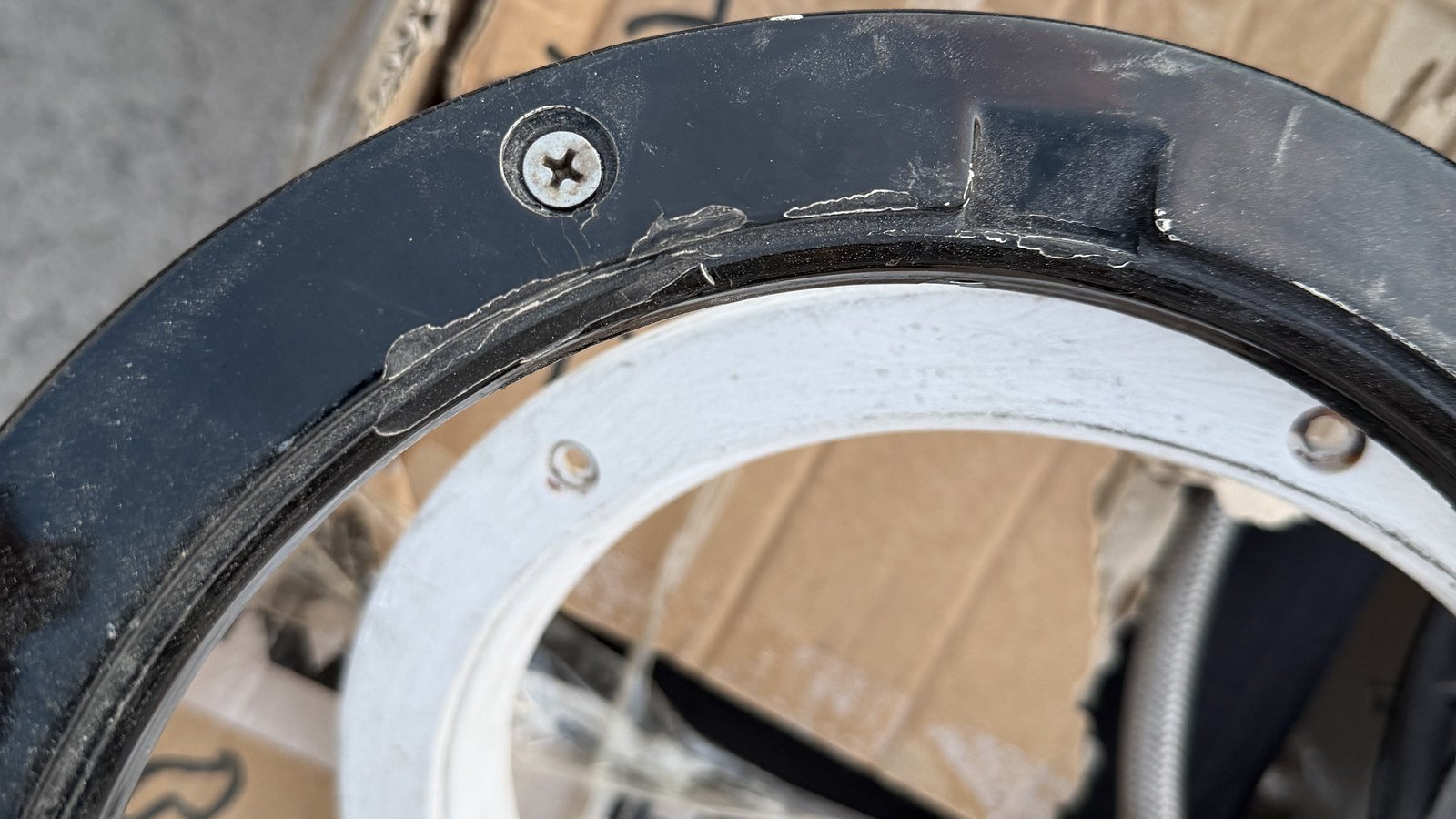 A removed curved black trim ring and its white mating ring flange, out of the vessel and lying on flattened cardboard cartons on a concrete floor, shot from directly above. Two small countersunk fixing holes are visible in the face of the white flange, one towards each side of the frame, one cross-head screw is still seated in a countersink in the black ring, and pale smears of old sealant run along the ring's inner edge in two or three places. Both rings run out of frame left and right, with more cartons and a grey braided flexible hose out of focus beyond.