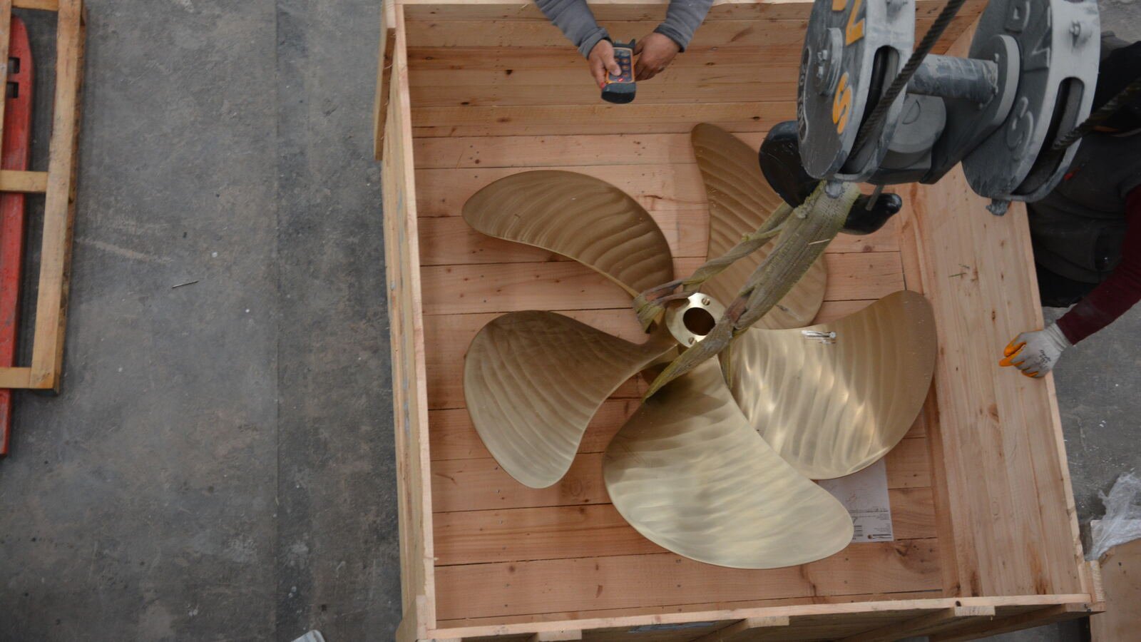A five-bladed bronze propeller lying in an open timber shipping crate, slung from a chain hoist while two pairs of hands steady it.