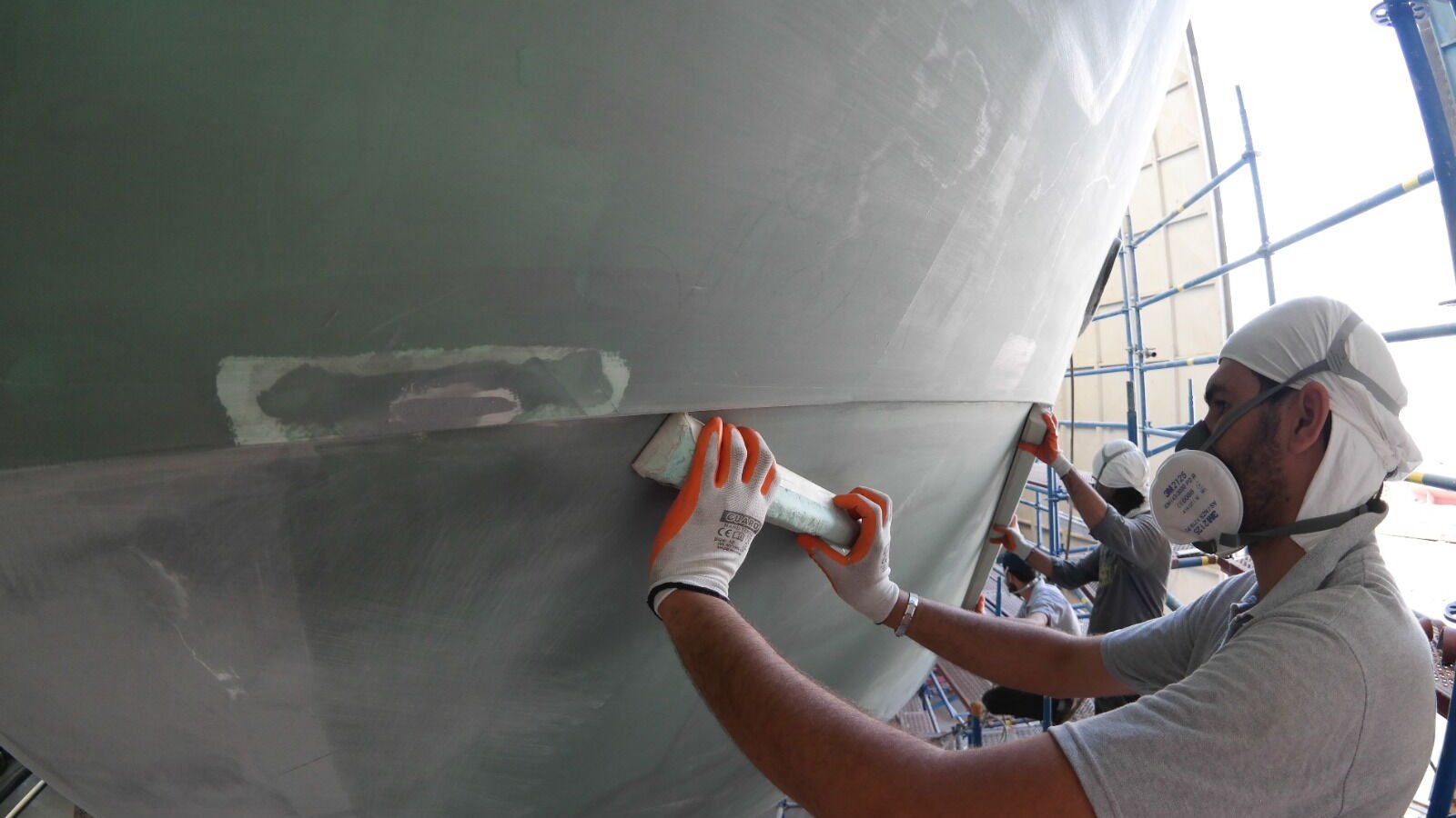 A worker in a cap and half-face respirator working filler by hand along a hull topside in pale grey primer, a feathered patch under his hands and scaffolding behind.
