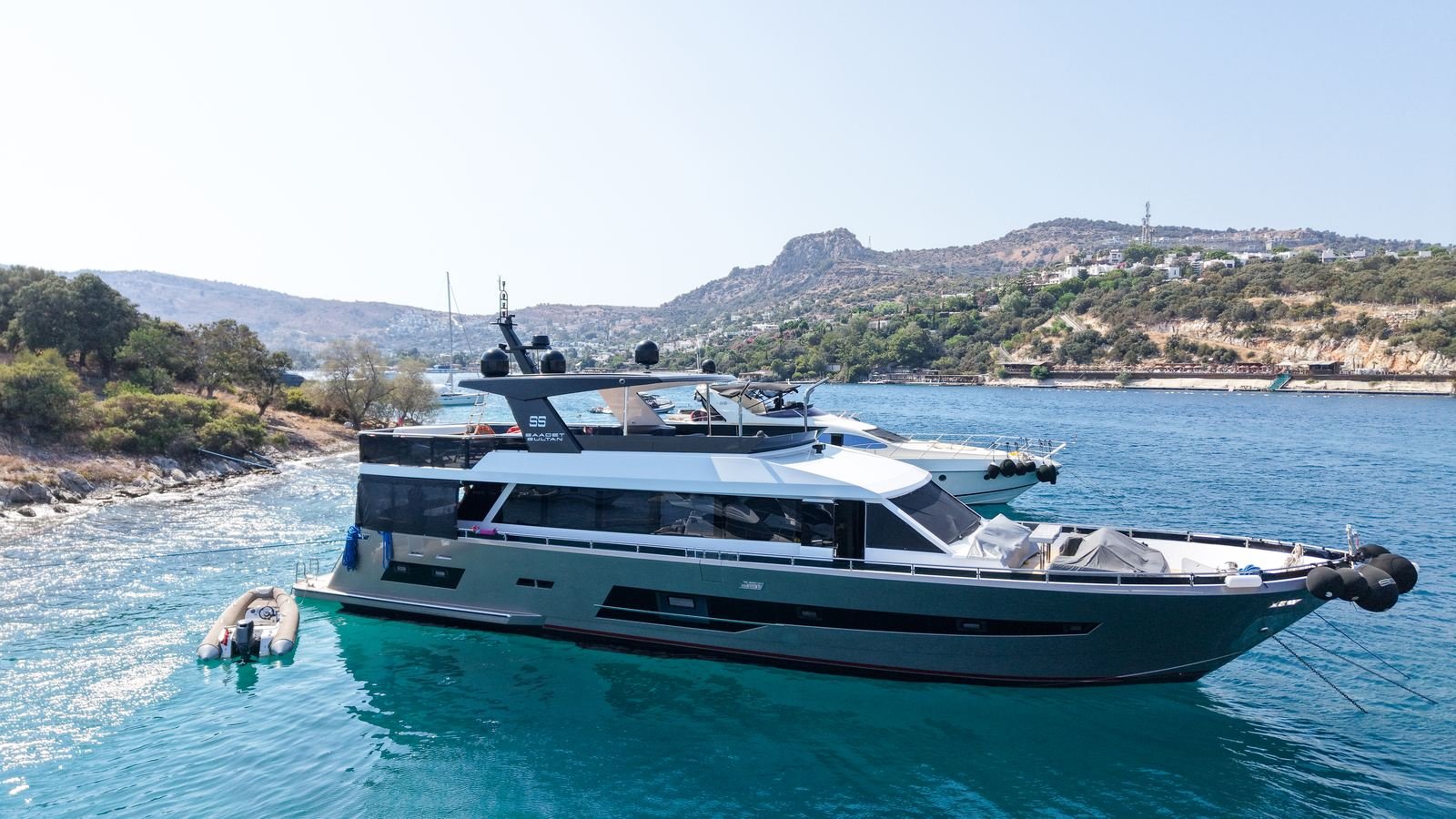 A motor yacht lying afloat in clear water off a rocky Aegean shoreline with her tender astern, seen from above after a refit