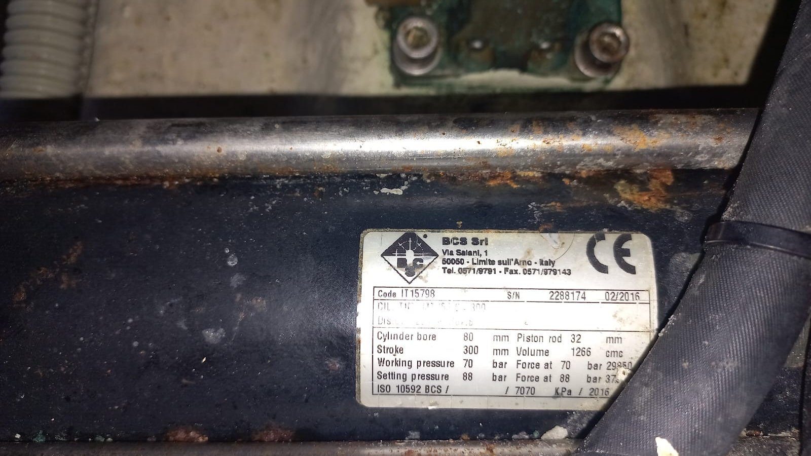 The stainless maker's plate on the barrel of a hydraulic cylinder, salt-crusted but still legible, on paint that is blistered and rust-streaked around it