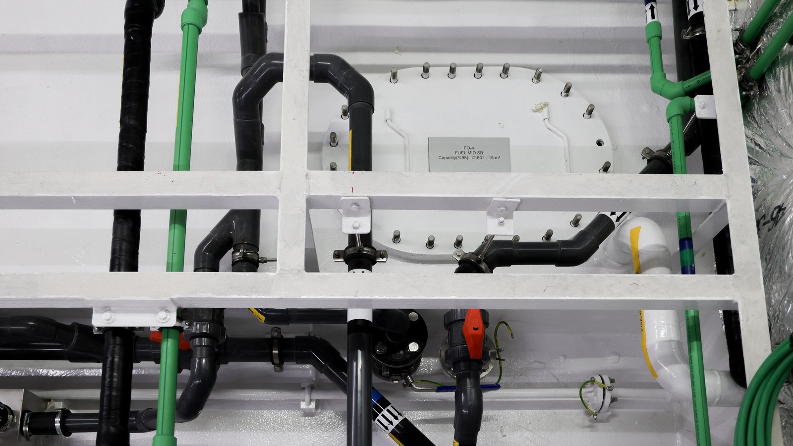 A small grey printed plate fixed at the centre of a white access cover in a tank face, its three printed lines giving a tank code, the fuel service the tank holds and a capacity figure. Short studs and nuts run round the edge of the cover, and a pale lifting bail lies against the white paint beside the plate. Green plastic pressure pipes rise on both sides of it, black pipes with swept elbows, clamps and a valve cross below, white-painted frame bars run horizontally across the foreground, and at the top edge of the frame a yellow adhesive tag is stuck along one pipe.