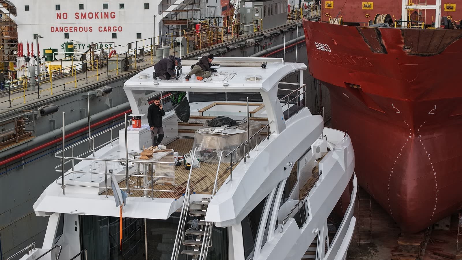 Three men working on the upper deck of a yacht in dry dock, joinery under protective sheeting, with the bow of a red commercial ship alongside