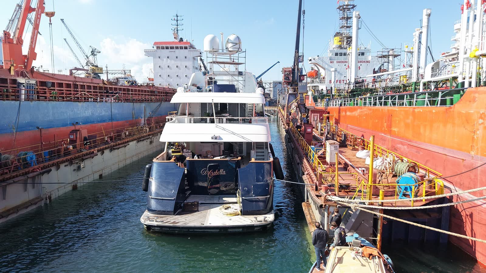 A dark-hulled motor yacht standing in a floating dock at Tuzla between two commercial ships, yard cranes above her and the repair district behind