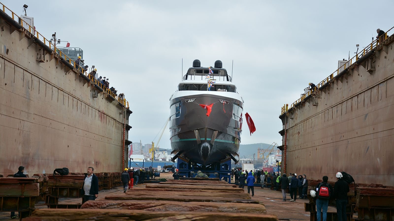 Bow-on view along the floor of a dry dock in Tuzla, rust-streaked steel dock walls rising on both sides with ladders and walkways along their tops, a dark-hulled motor yacht standing on keel blocks at the far end with timber shores under her bilges and a Turkish flag at her bow, and some forty yard workers in hard hats and high-visibility jackets spread across the timber-strewn dock floor.