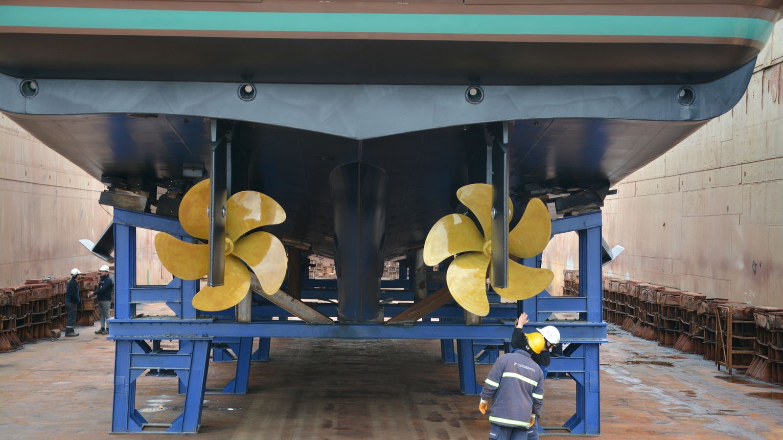 A motor yacht chocked in a dry dock with her twin five-bladed propellers and shafts exposed, a yard hand standing beneath the transom for scale