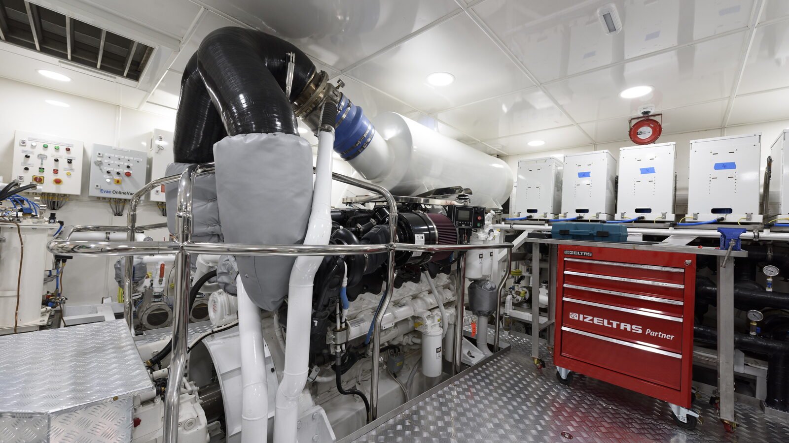 A finished machinery space aboard a yacht, lit and in commission, looking along one of the main engines. A black corrugated exhaust bellows rises from the engine in a tall arc to a stainless clamp and a blue silicone joint, and a white lagged exhaust silencer runs away behind it. A grey quilted removable insulation blanket is laced over the turbocharger and exhaust elbow below the bellows. A polished stainless guard rail runs round the engine at waist height. The engine itself is painted white, with a conical red air filter element on its induction side, black hoses, spin-on filters and blue-sheathed small-bore lines along its flank. Aluminium chequer plate covers the deck and a chequer-plate box stands over the gearbox in the left foreground. On the right, a red five-drawer rolling tool chest lettered IZELTAS Partner stands on the chequer plate against a stainless-topped bench, with a blue plastic case on the bench above it, and beyond the bench five tall white equipment cabinets run along the ship's side, each marked with a short strip of blue tape. A round red fire detector is fixed to the deckhead on that side. Two small white control panels are mounted on the bulkhead at the far end, one of them lettered Evac OnlineVac. The deckhead is a white panelled grid with recessed downlights and a louvred supply grille at the upper left corner. Nobody is in the frame.