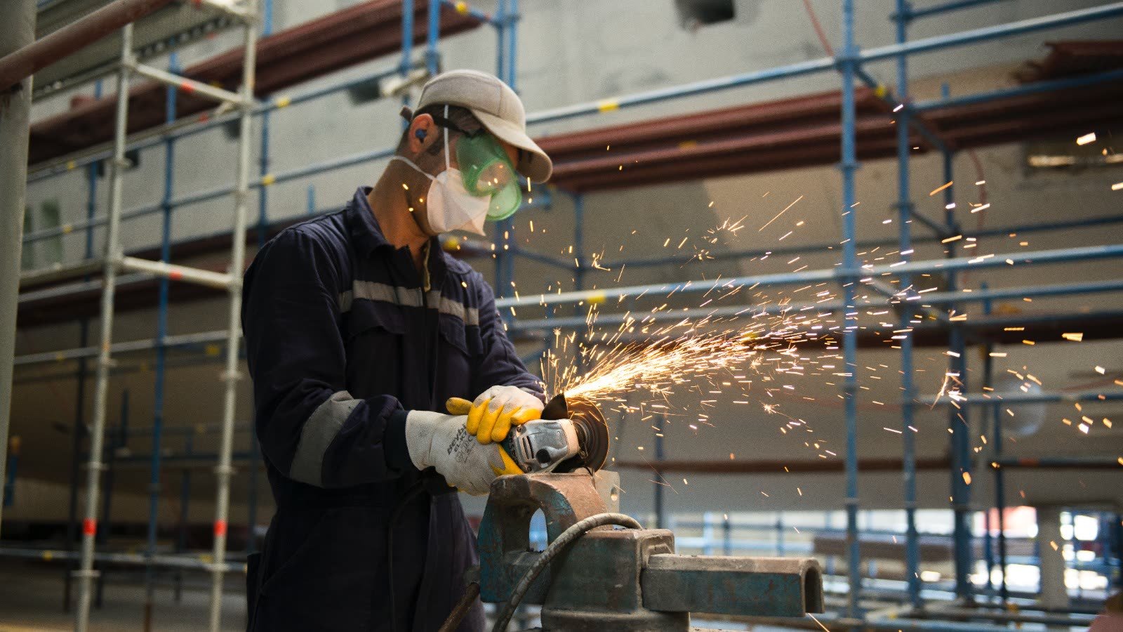 A yard hand in overalls and a respirator grinding a steel part at a bench inside a covered refit hall in Tuzla, sparks arcing past the scaffolding