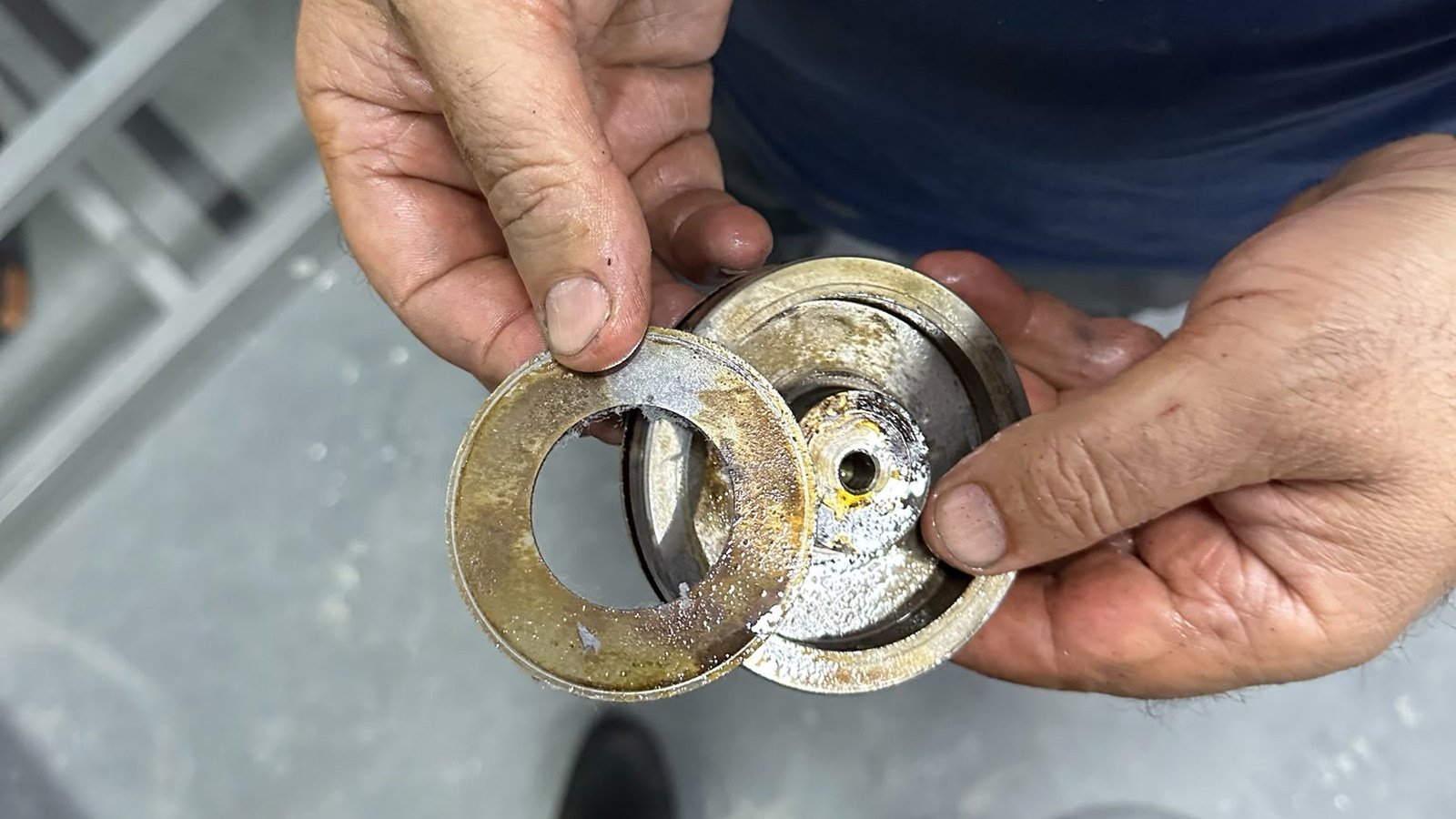 Shot down over a man's shoulder in flat, even light as he holds two removed components up towards the camera, one in each hand and overlapping. Nearer the camera, in his left hand, is a flat ring with a plain round bore through it, its face mottled yellow-brown and speckled with pale crystalline deposits, and a broad patch of pale grey bare metal near his thumb where the mottling gives way. Through the bore, the curved edge of the component behind shows, part bright and part in shadow. Behind it, in his right hand, is a deeper stepped fitting whose machined surfaces are bright and pale-speckled, with yellow staining around a small drilled hole set off-centre. A navy torso fills the upper right. A grey floor runs below, with a dark boot at the bottom edge, and at the upper left pale bars cross the frame, too far out of focus to name. He is holding one part up against the other.
