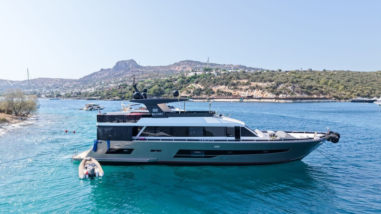 A motor yacht lying at anchor in a sheltered bay, her tender made fast on a line astern, with scrub-covered hills and white houses on the shore behind her