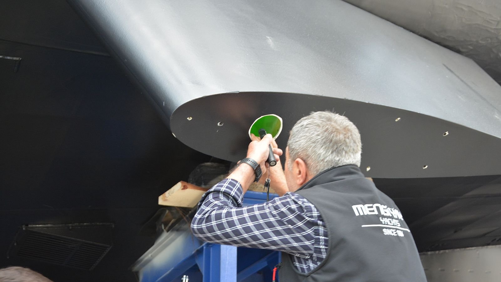 Seen from below, a grey-haired fitter in a Mengi Yay Yachts gilet and checked shirt reaches up from a blue staging tower with a green-shrouded handheld tool to work on the underside of a dark hull where a stabiliser fin meets the plating, a line of fastener holes running along the fin root.