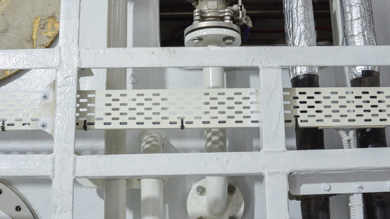 White-painted pipework, bolted flanges and a valve body running through the frames of a yacht machinery space
