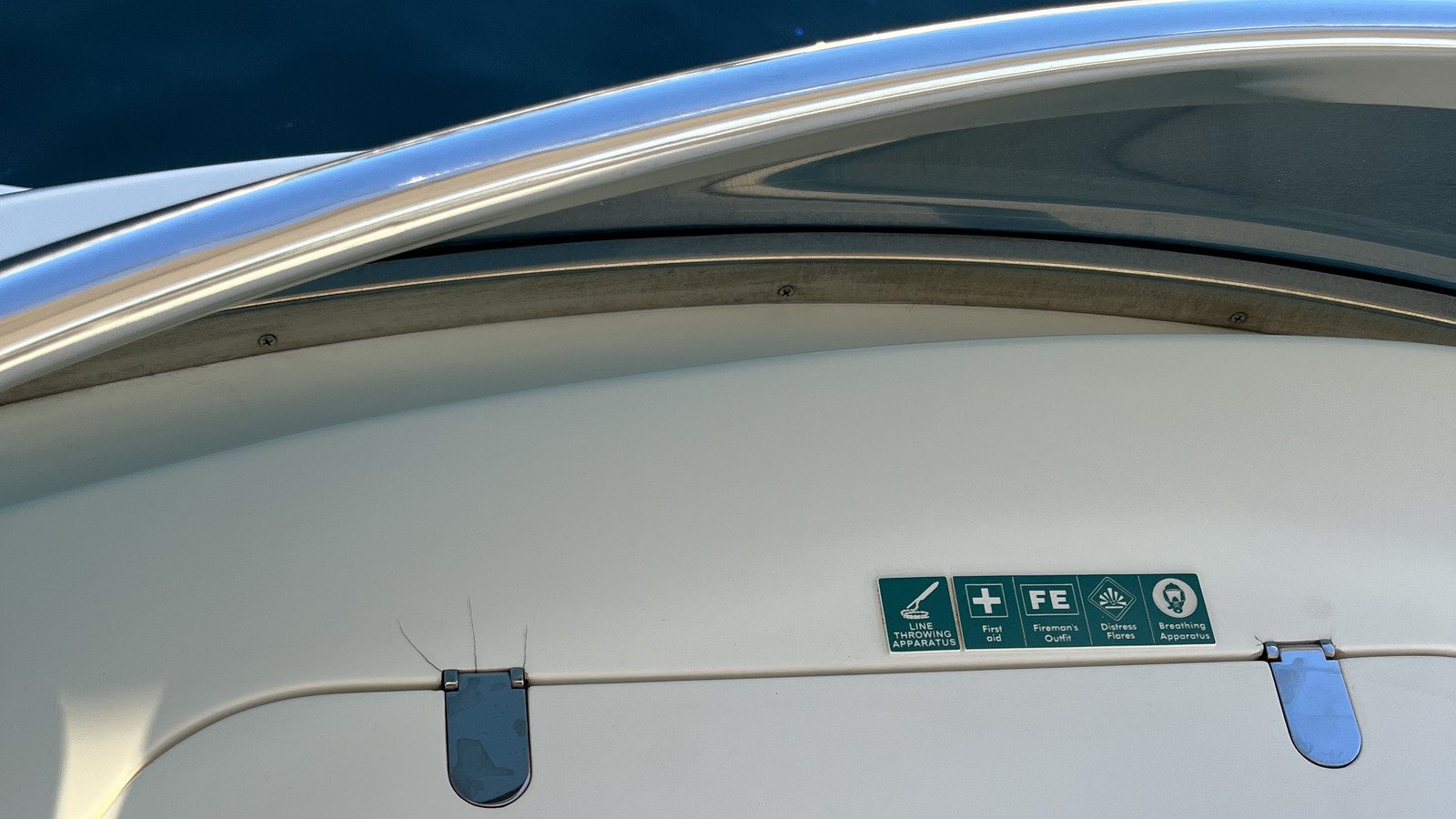 A green and white sign board fixed to the plain white side of a deckhouse, its five tiles reading in order LINE THROWING APPARATUS, First aid, FE Fireman's Outfit, Distress Flares and Breathing Apparatus, each set with its own white pictogram: a line-throwing device, a cross, boxed capitals, a radiating starburst and a breathing mask. The board sits just above a fine horizontal joint running the width of the frame, with a dark grey rounded clip straddling that joint to the left of the board and a second near the right edge; a few fine pale fibres are caught under the left clip. Above, a mirror-polished stainless handrail sweeps across the upper third in a shallow arc and throws a hard specular highlight along its crown, with a weathered grey-gold timber capping below it carrying three countersunk screw heads. Beyond the rail at the upper left is a flat dark navy surface, and to its right a large dark tinted pane with a soft greenish-bronze reflection across it. The white paint is clean and unmarked. None of the equipment the board names is visible in the frame.