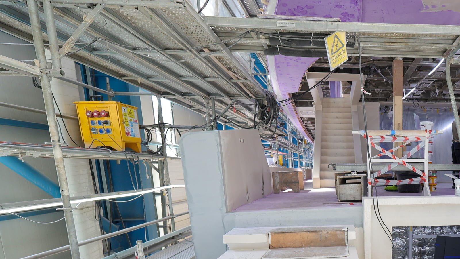 Inside a covered refit hall at Tuzla, looking along a deck level of a motor yacht under refit: at left a yellow temporary site distribution board bolted to a scaffold tube at chest height, with three red three-phase sockets in a row above four blue single-phase sockets, Turkish warning placards on its front, and black temporary cables looping away along the scaffold tubes and hanging coiled overhead. A yellow diamond hazard sign reading Dikkat! above Alcak Tavan hangs from the staging in the centre. Scaffold towers and perforated staging boards run along the vessel's side with LED strip lights cable-tied to them, and beyond are a primed white bulwark, a deck coated in lilac primer and blue shed cladding.