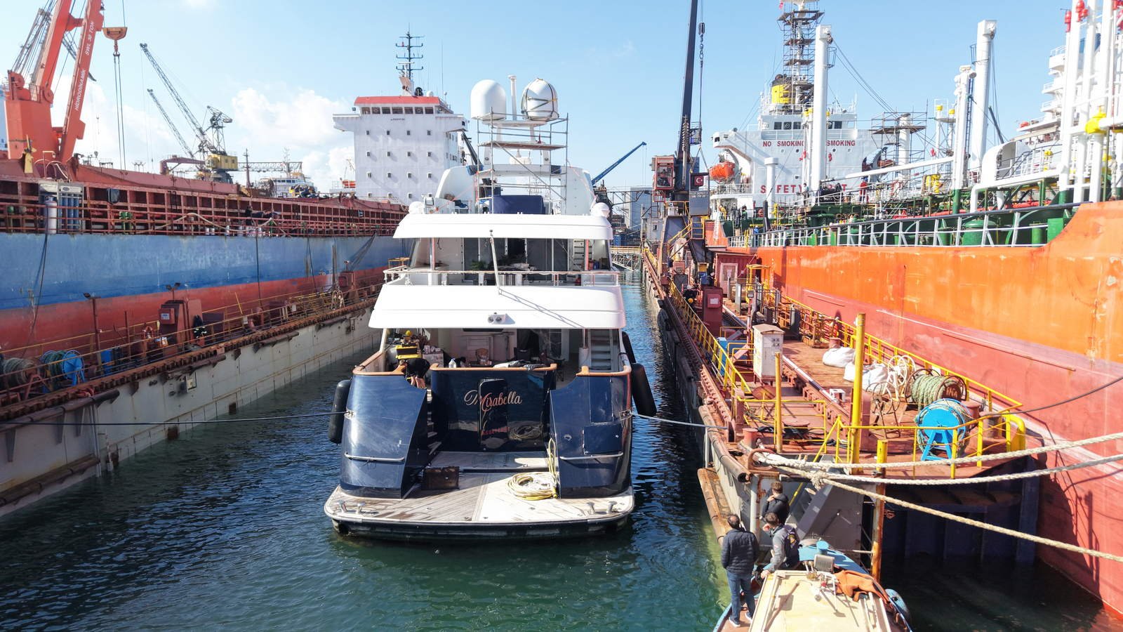 A superyacht lying in a floating dry dock at Tuzla between the plated sides of a bulk carrier and a tanker, shipyard cranes and staging standing over the basin