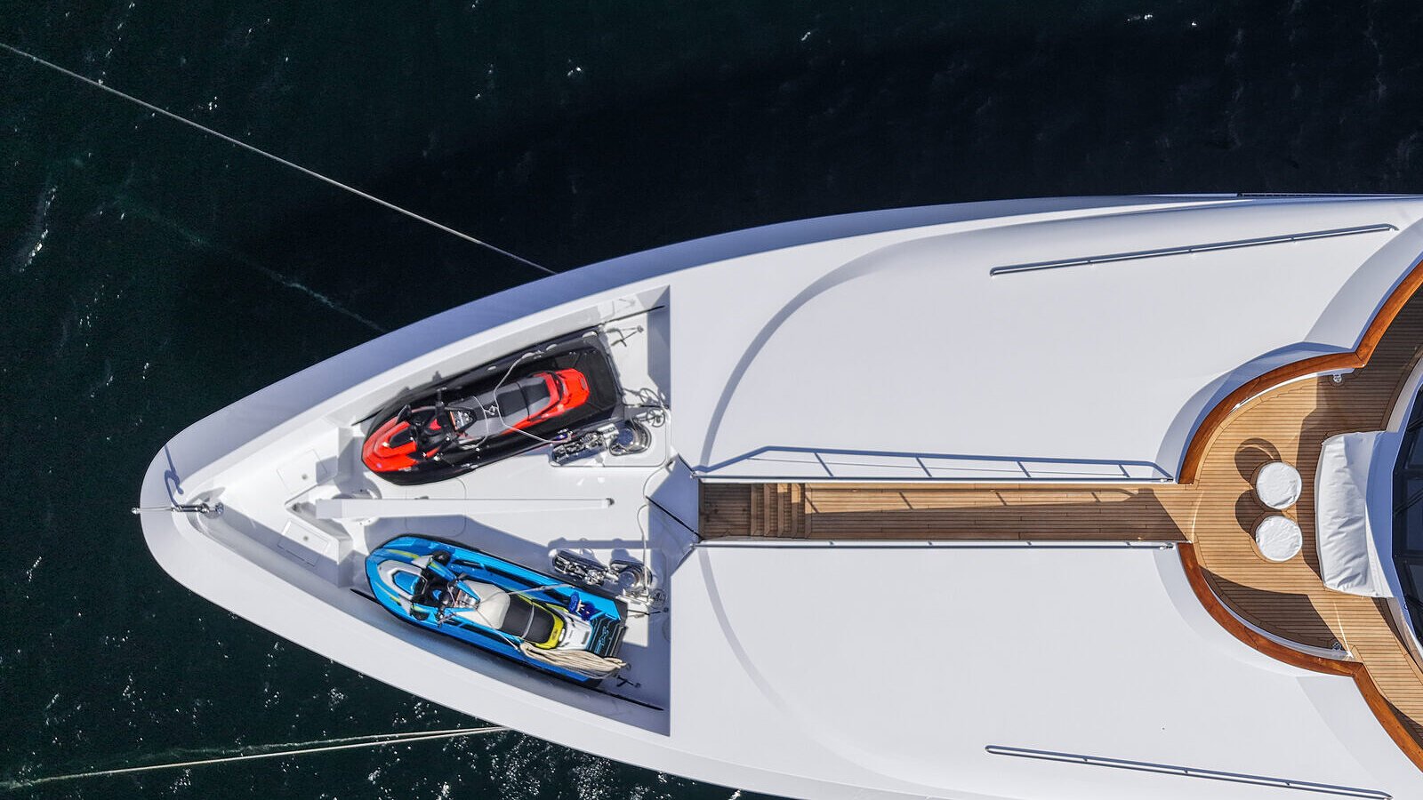 A straight-down aerial of a bow, two personal watercraft side by side in a recessed foredeck well, each sitting in a fitted cradle with strap tie-downs.