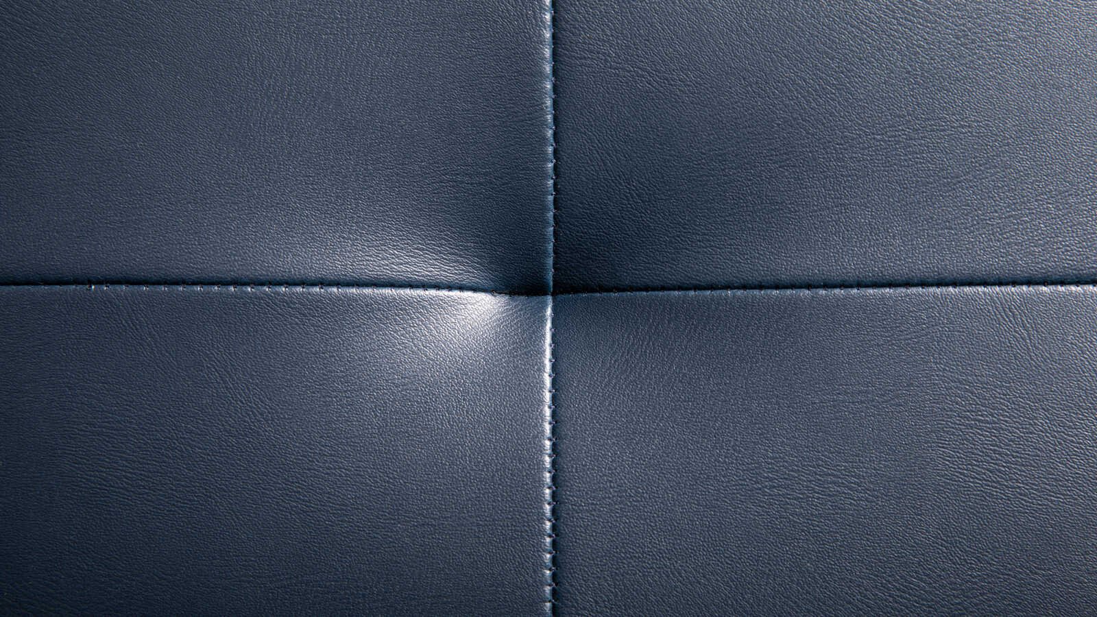 A close view of padded navy leather, four panels meeting at a cross of two stitched seams, the leather rolling slightly proud either side of each line of stitching.