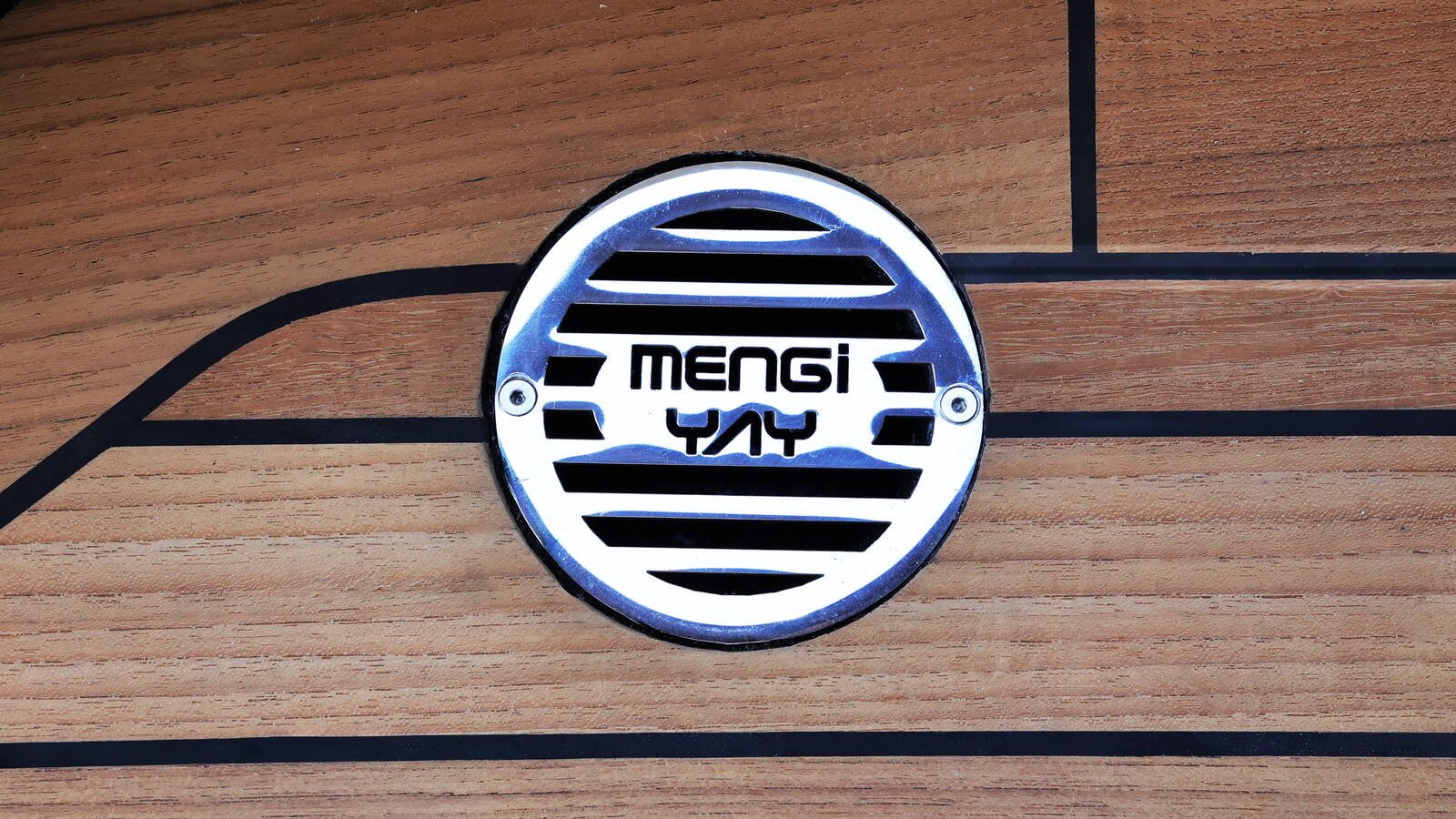 A round louvred fitting let into a teak deck, seen from directly above and filling the middle of the frame. The fitting is a polished stainless disc with horizontal slots cut right through it, the slots shortening towards the top and bottom edges to follow the circle, and the words MENGI YAY cut through the two broad slots across its middle. Two small countersunk screws sit in the rim, one on each side at the level of the lettering. The disc is bedded on a ring of black mastic that shows all the way round its edge and is widest at the lower left. The teak runs diagonally across the frame from lower left to upper right, three or four boards legible, with straight black caulk seams between them; one seam curves away round the lower left of the fitting as the edge of a margin board, and another runs square across the upper right corner. Nothing below the deck is visible through the slots, which read as flat black.