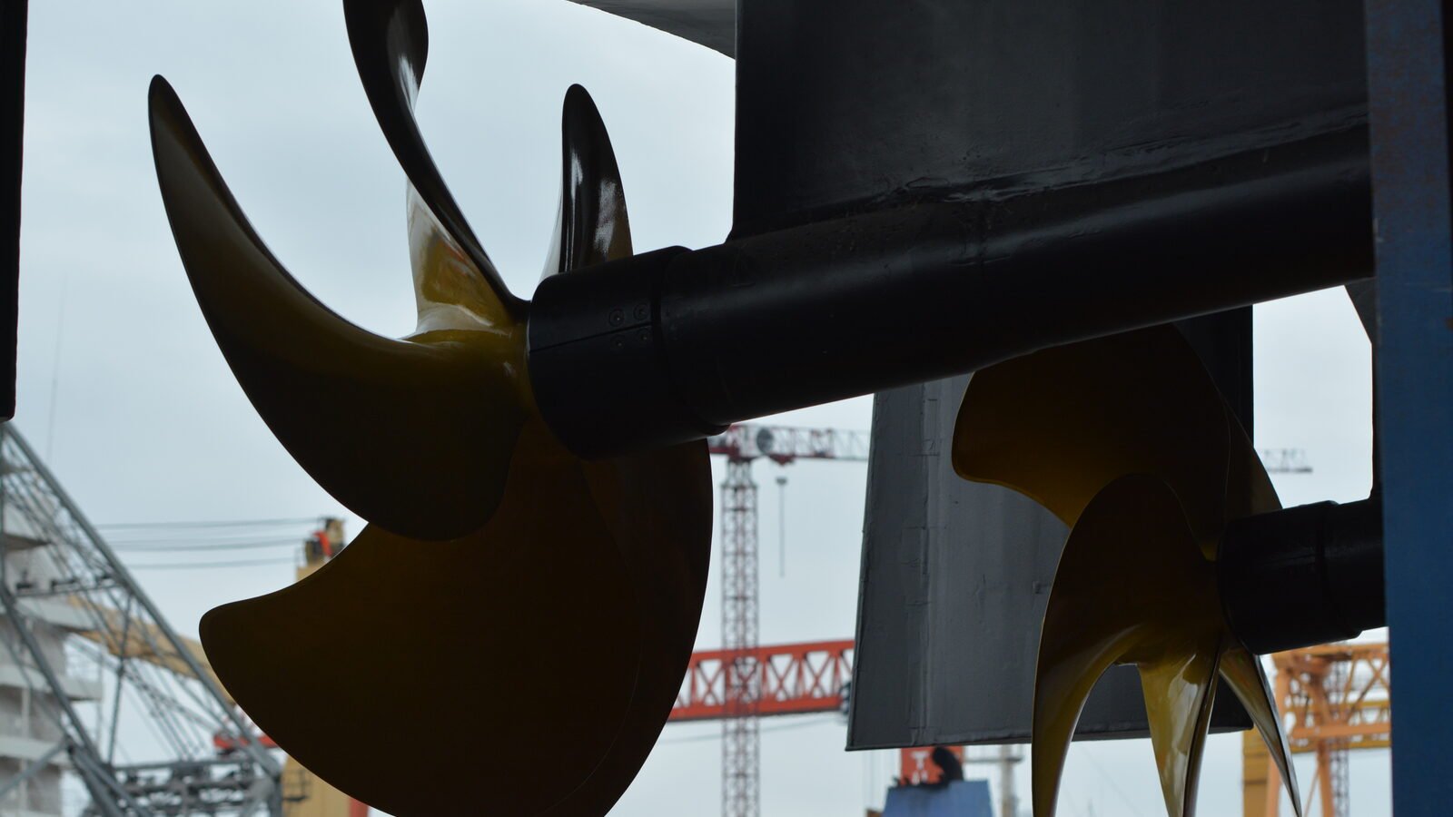 A yacht's propeller and rudder under the hull ashore in the yard, shipyard cranes standing beyond