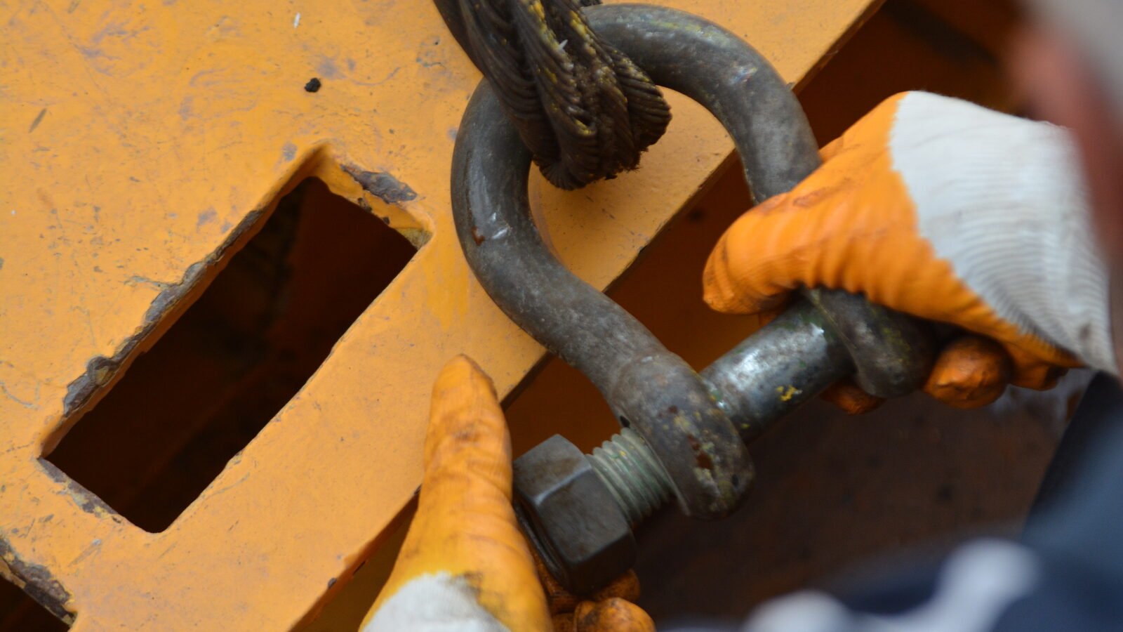 Gloved hands making up a galvanised shackle through a slot in a yellow-painted steel fitting