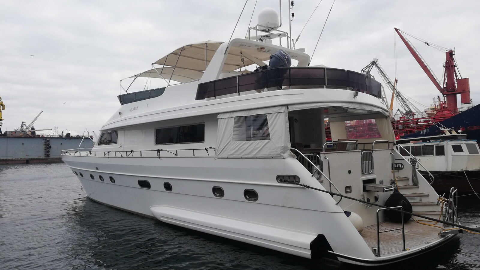 A motor yacht lying alongside in a working shipyard under flat grey cloud, yard cranes and commercial berths behind her
